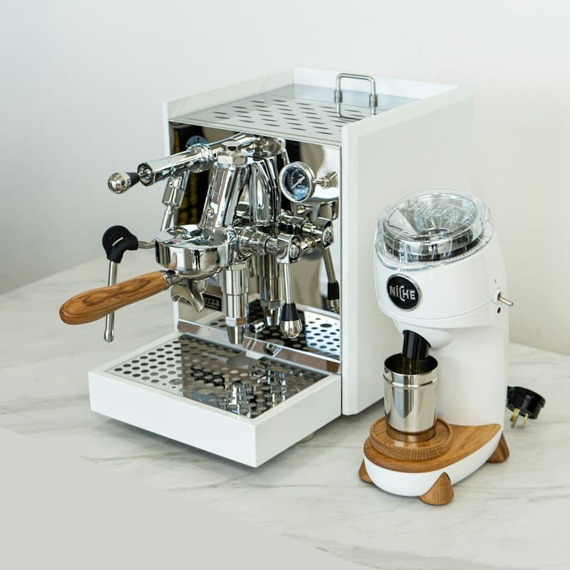 A white and chrome espresso machine with a wood-handled portafilter next to a white grinder on a pale marble counter
