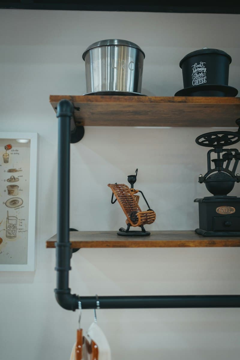 Two wood shelves on black pipe brackets holding metal coffee filters, a vintage hand grinder, and a small decorative figure