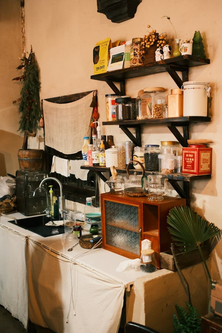 Open wall shelves above a kitchen counter with a small sink, holding jars of coffee and tea, canisters, and glass teapots