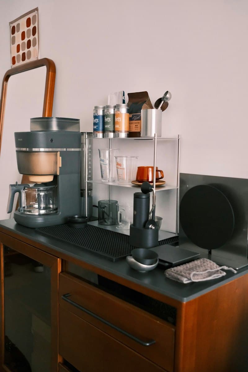 A drip coffee maker, hand grinder and small steel shelf of cups and coffee tins on a dark countertop over a wooden cabinet