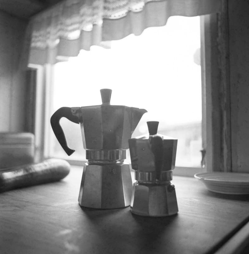 Two aluminum moka pots, one large and one small, on a wooden table in front of a bright window