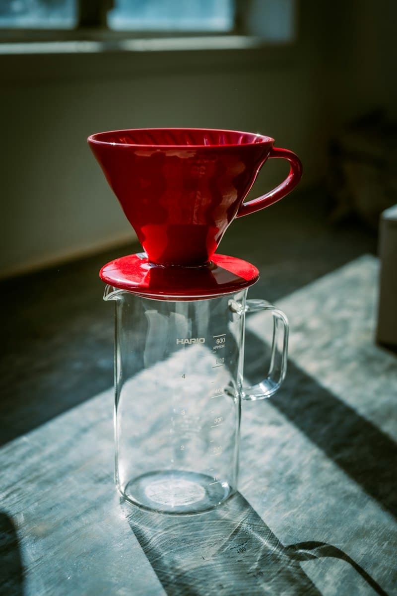 Red ceramic pour-over dripper sitting on a glass measuring server on a sunlit wooden table