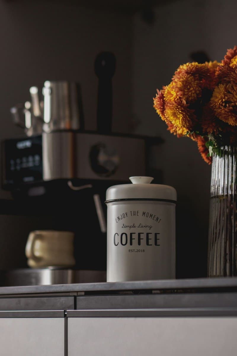 A white metal coffee canister and a glass vase of orange flowers on a counter, with an espresso machine out of focus behind them